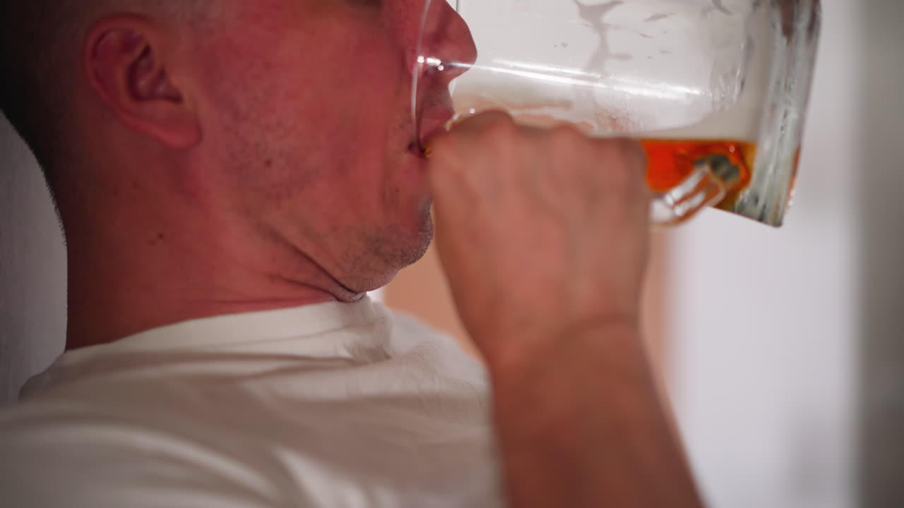 Close up of white man leaning against wall wearing white polo drinking berry wine from transparent glass with soft blurred indoor background