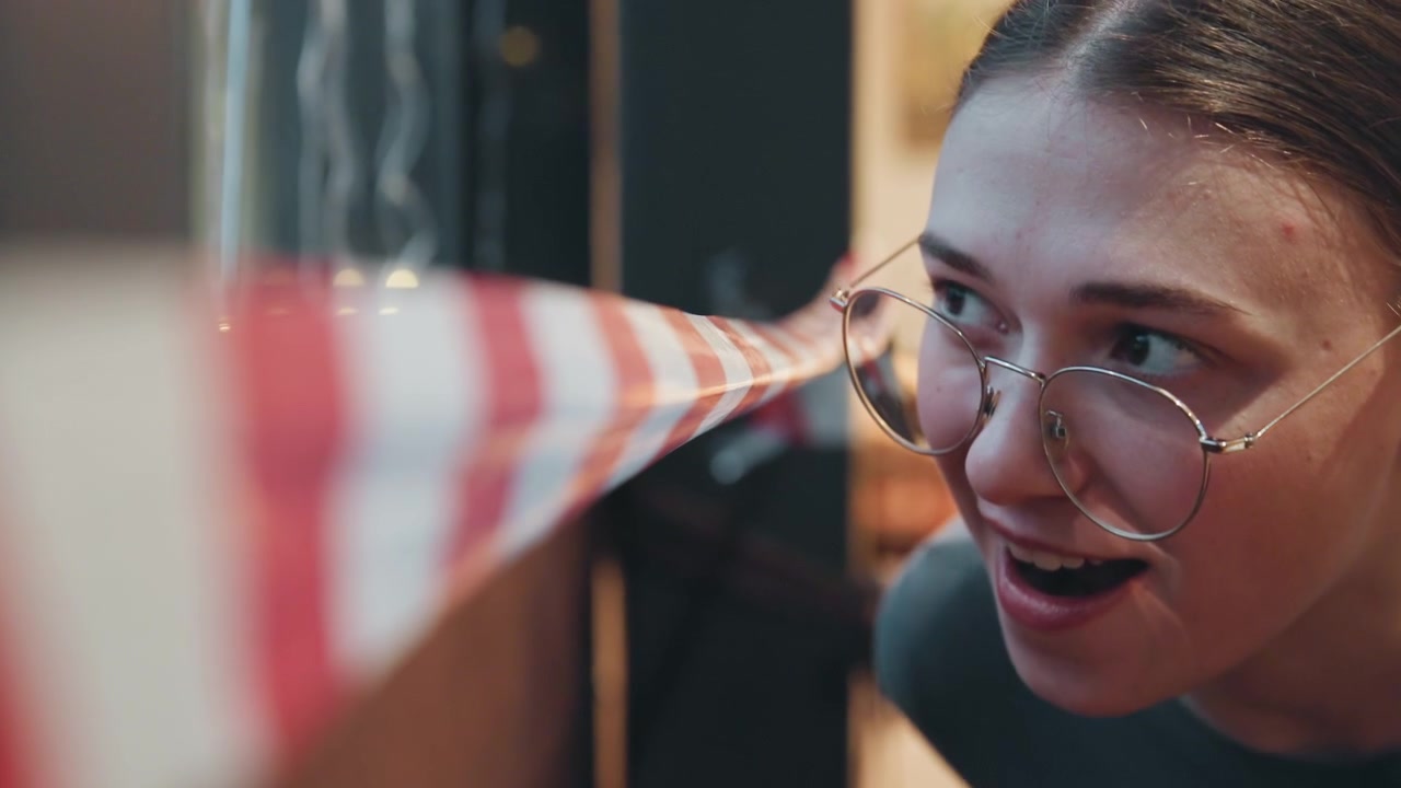 Caucasian woman peeking past caution tape, glasses closeup in dim cafe interior, leaning over redwhite barrier, inquisitive expression, evening bokeh lights and vintage film grain