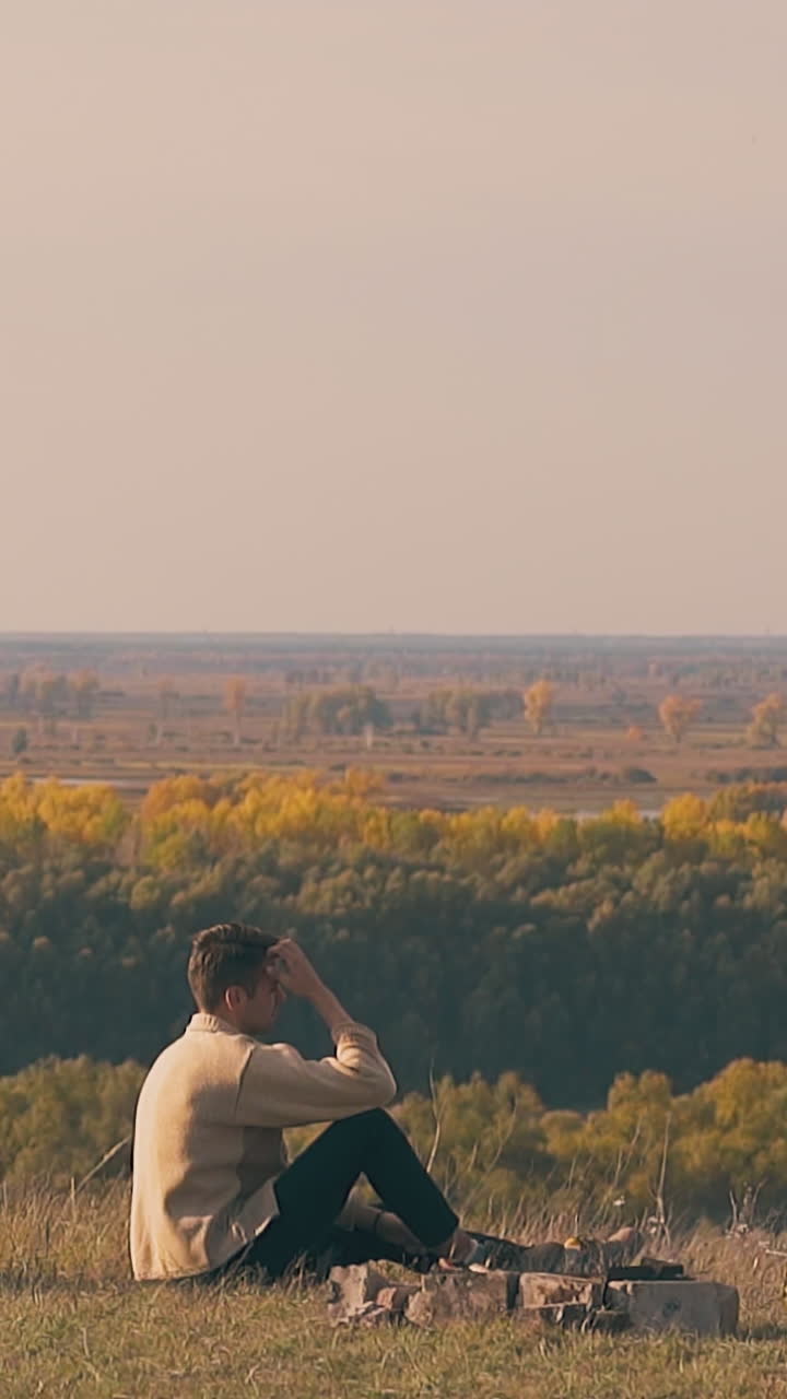 sad guy sits by camp bonfire on river bank and looks at green and yellow autumn forests in early morning slow motion