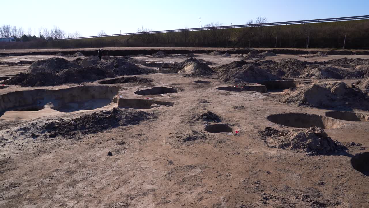 Push in through archaeology site past holes, mounds of dirt. Highway in background
