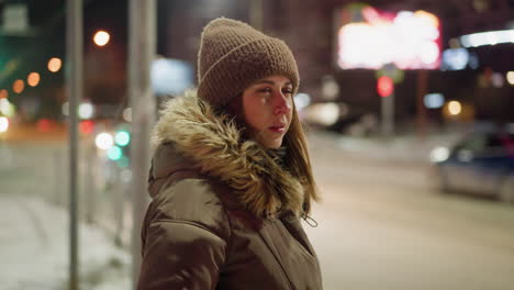 first-person view of a woman in a winter coat with a fur hood, standing and rubbing her hands together while looking to the right at night. blurry cars are moving in the background