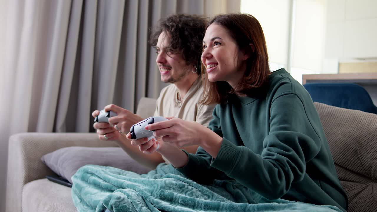 feliz chica morena junto con su novio con el cabello rizado envuelto con una manta y jugando videojuegos mientras está sentado en el sofá usando un joystick en casa