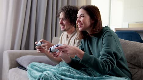 Chica-Morena-Feliz-Junto-Con-Su-Novio-Con-Cabello-Rizado-Envuelto-En-Una-Manta-Y-Jugando-Videojuegos-Mientras-Están-Sentados-En-El-Sofá-Usando-Un-Joystick-En-Casa