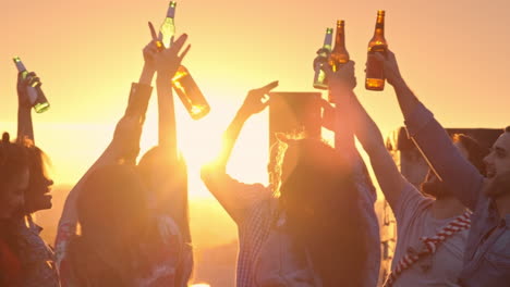 Group-Of-Friends-Enjoying-A-Party-On-A-Terrace-At-Sunset,-Hold-Drinks-With-Arms-Raised