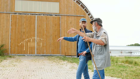 side view of old father and son farmers walking and talking outside the stable