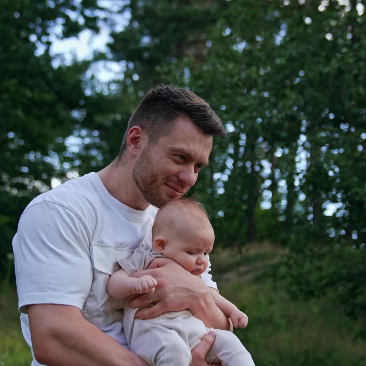 Man waving his baby boy in hands. Caring happy dad kisses child's head. Blurred nature at backdrop
