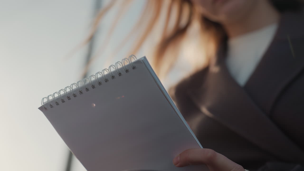Woman holding spiral notebook closeup, reporter jotting notes during interview on city street, warm backlight rim highlighting hair, shallow depth of field, professional coat, subtle hand movements
