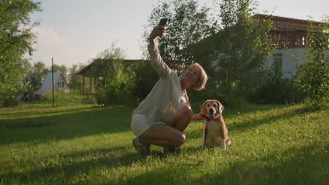 woman taking selfie with her happy dog outdoors in lush green garden under sunny weather, with building and trees in background, dog is sitting, panting joyfully, as owner holds phone smiling