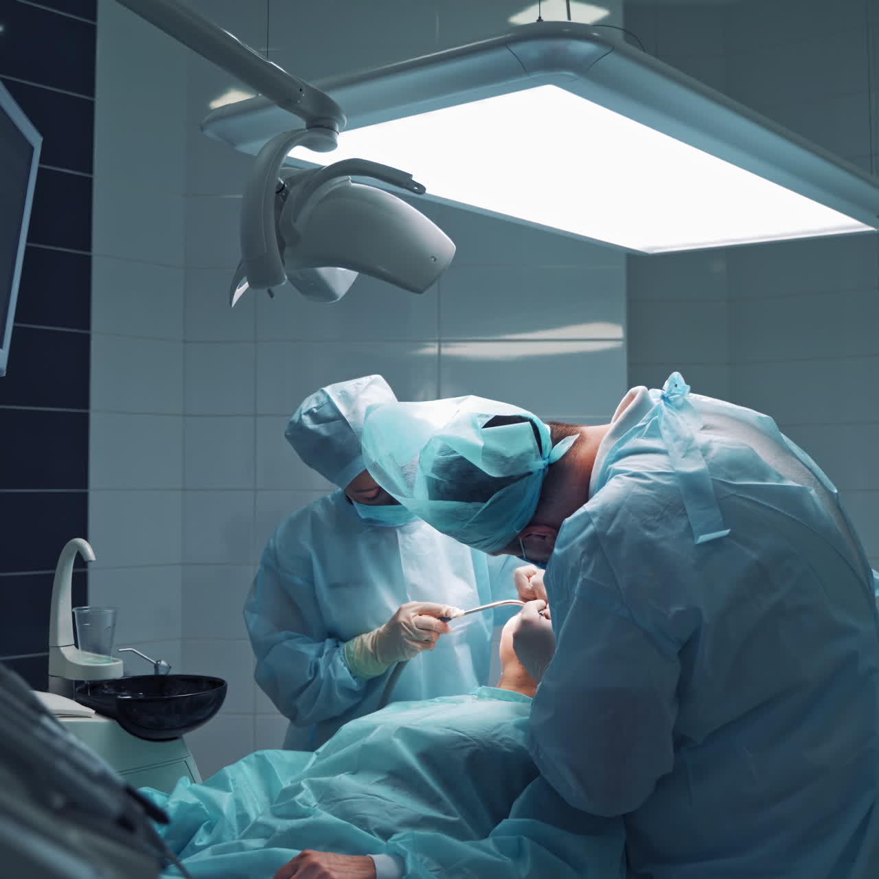 Doctors in medical masks are doing tooth treatment in modern stomatology clinic. Male professional dentist with his assistant at work. Operation procedure.