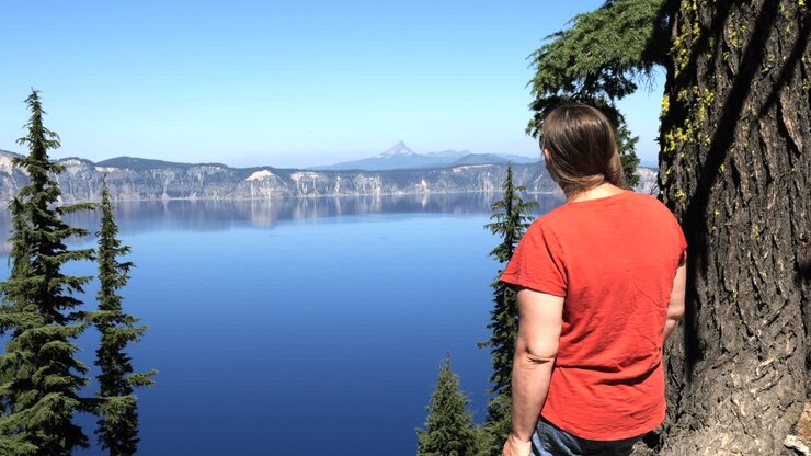Donna in piedi accanto a un albero che si affaccia sul Crater Lake con una cima montuosa in lontananza