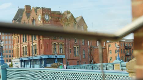 pedestrians cross ornate city bridge, red brick buildings in background, daylight, slight camera movement
