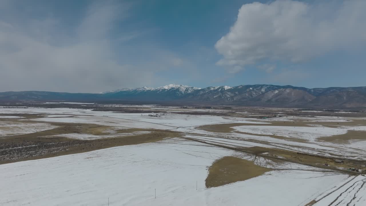 Bredt luftlandskab af snedækkede marker og frossen steppe med en fjern bjergkæde under en skyet blå himmel, vinterlandskab i Buryatien, Rusland