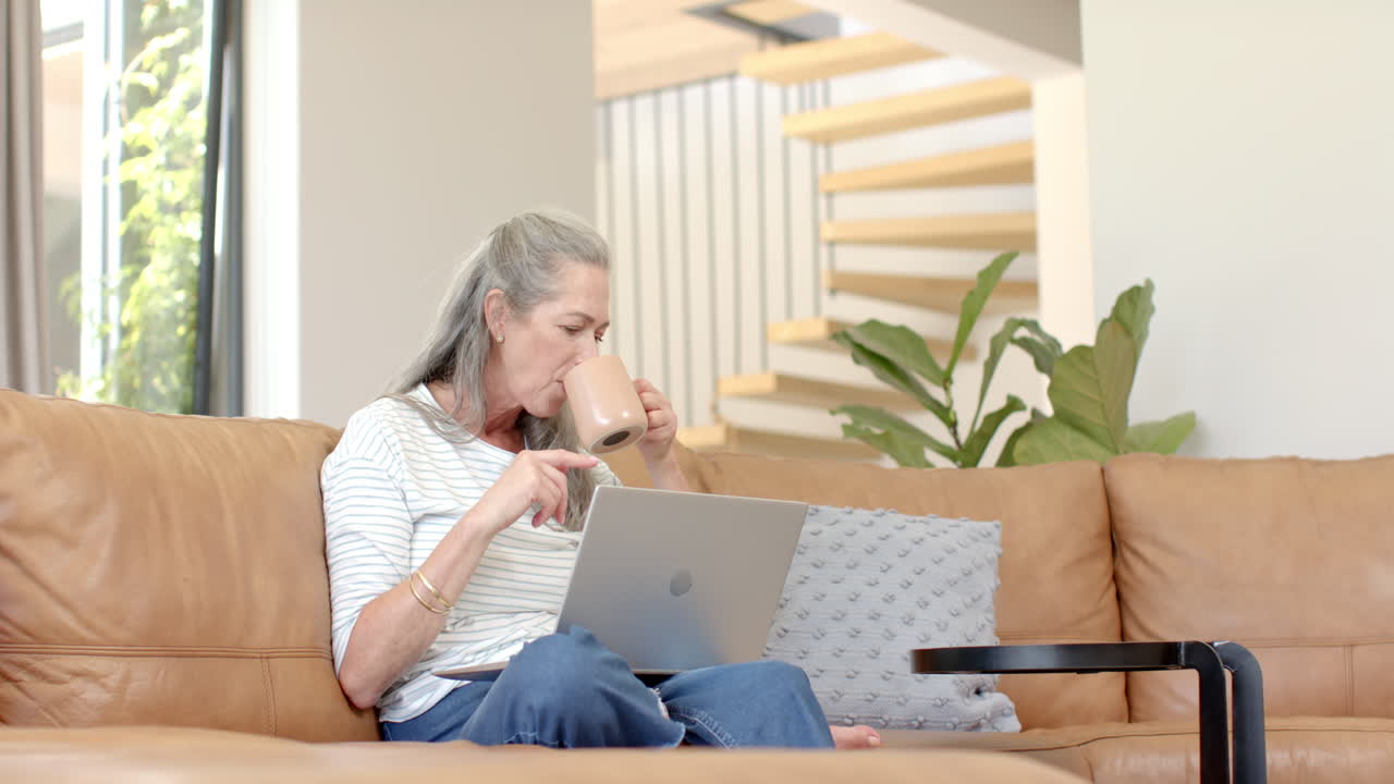 Mature Caucasian woman sitting on sofa, drinking coffee and looking at laptop