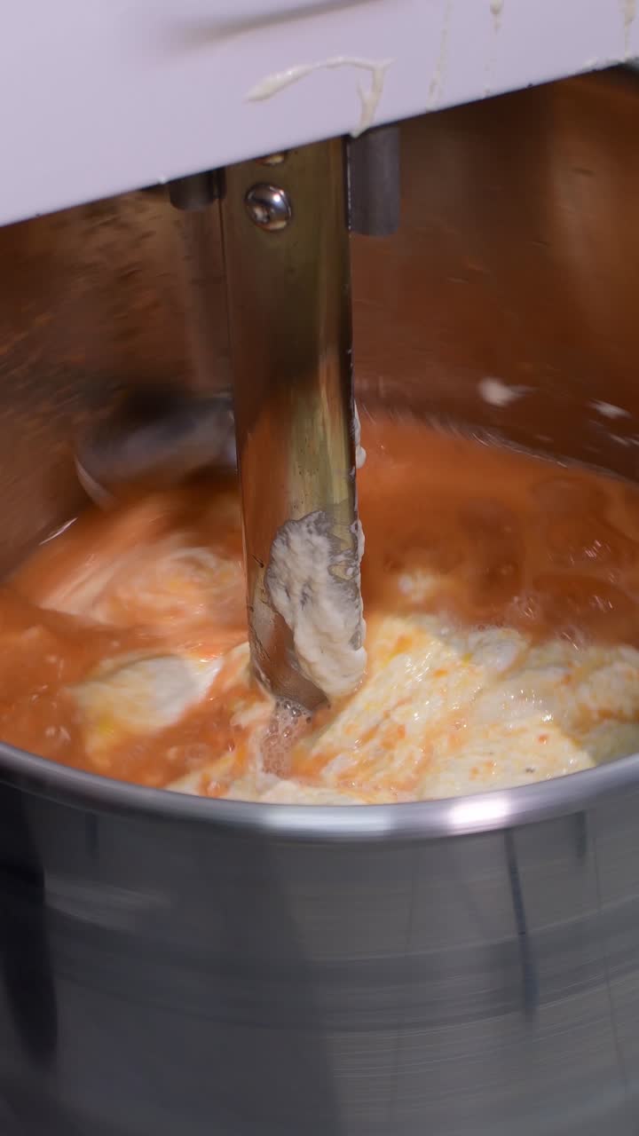 Primo piano di un'impastatrice industriale che aggiunge uova all'impasto mentre un braccio miscelatore rotante amalgama gli ingredienti in una massa liscia ed elastica. Impastatura meccanica, trasformazione alimentare commerciale, panificio su larga scala.