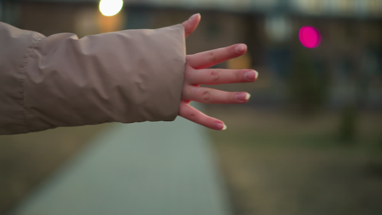 un primer plano captura la mano de una persona en una chaqueta de melocotón, con los dedos extendidos, en un fondo urbano borroso con iluminación suave y efectos bokeh