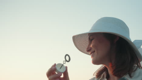 young woman wearing white sun hat blowing delicate soap bubble into glowing sunset sky framed by winding river valley and tall grassy hillside