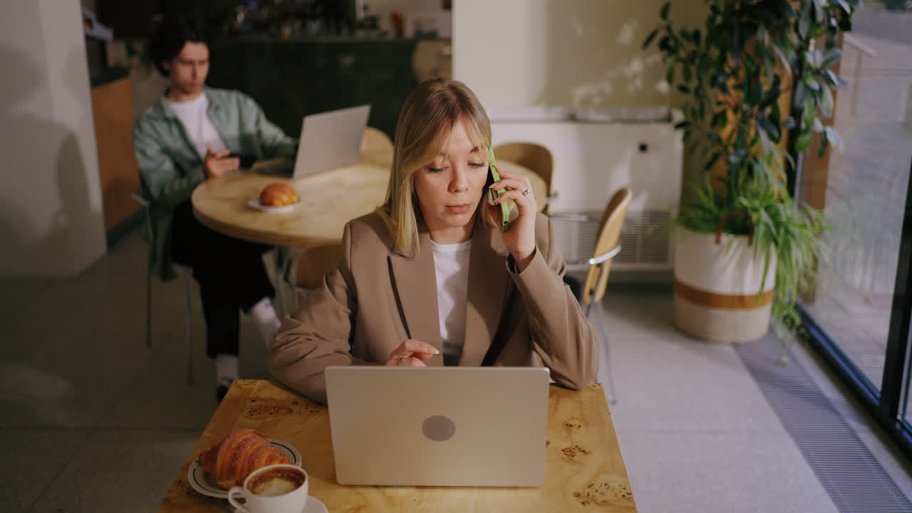A woman on a phone call in a coffee shop, working on her laptop.
