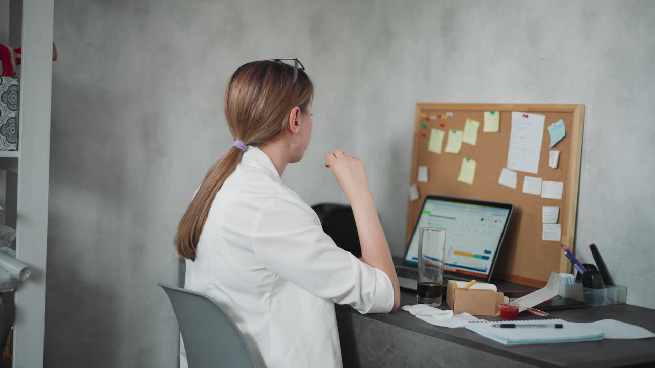 Young woman with hair tied back wearing white shirt sitting at desk during break eating fries dipped in red sauce while working on laptop in cozy home office space with cork board filled with notes