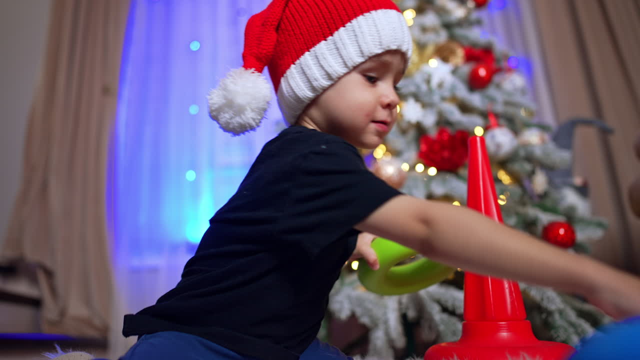 Cute Caucasian toddler in red cap building a pyramid. Christmas tree decorated for holiday at backdrop. Low angle view.