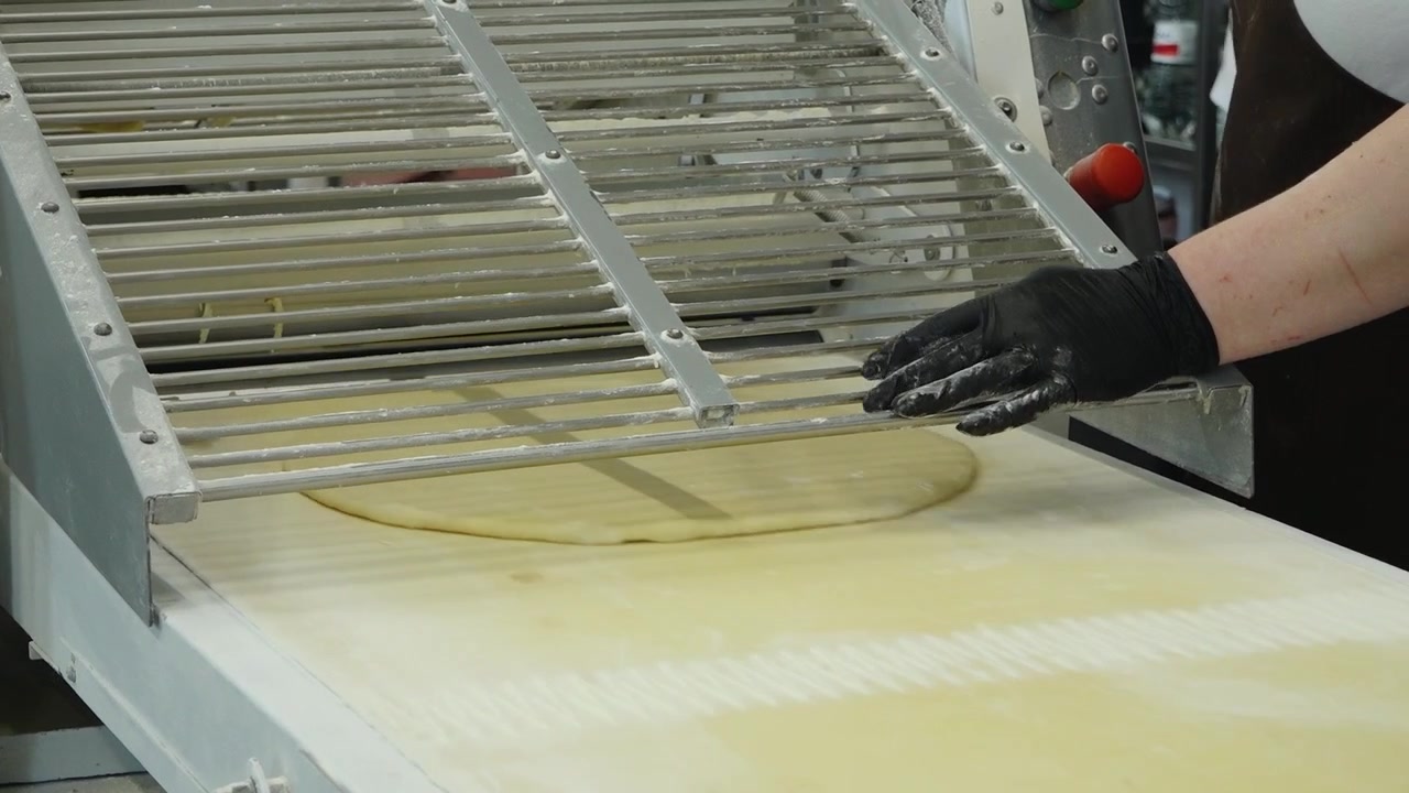 A gloved baker works with a flattened dough sheet passing through industrial bakery sheeting equipment during pastry preparation.