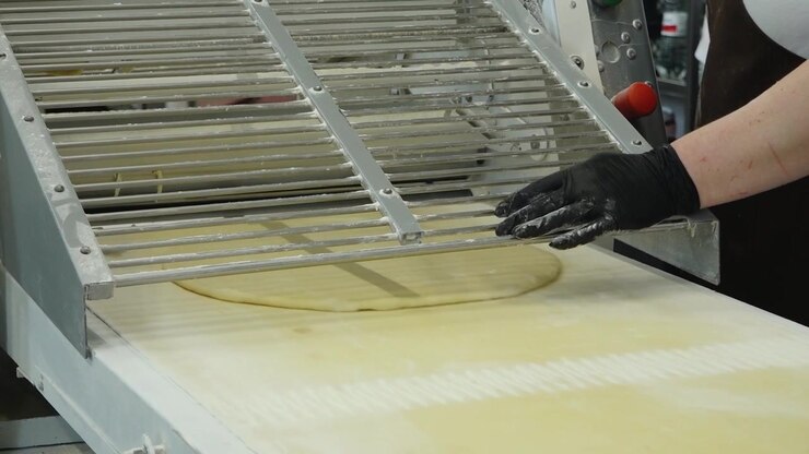 A gloved baker works with a flattened dough sheet passing through industrial bakery sheeting equipment during pastry preparation.
