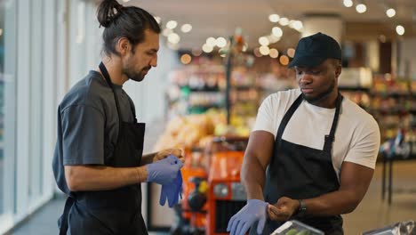 A-man-with-Black-skin-and-a-beard-in-a-white-T-shirt-and-a-black-apron-communicates-with-his-colleague-a-brunette-man-with-a-beard-in-a-gray-T-shirt-during-their-duty-at-a-grocery-store-counter.-And-male-store-workers-wear-protective-gloves