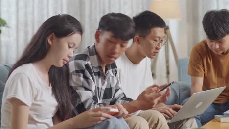 close up of asian teen group studying at home. students with books and laptop looking at smartphone, celebrating success in project, giving high five gesture