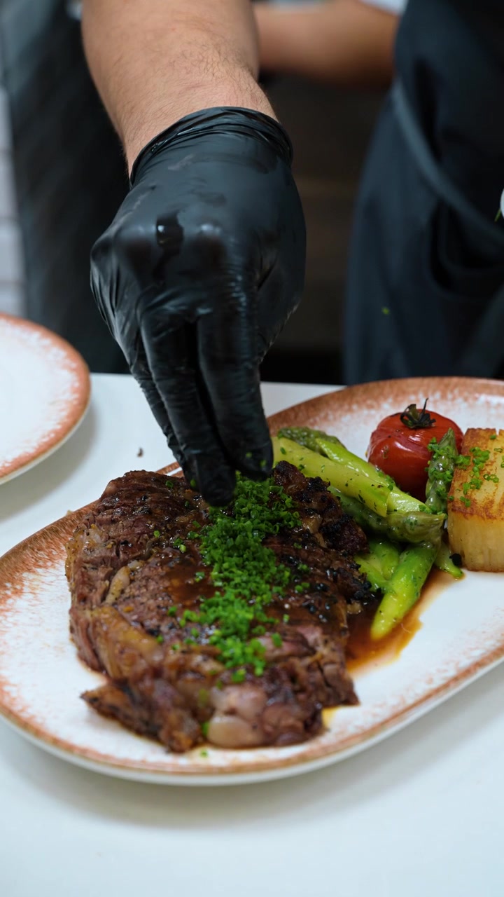 Chef garnishing grilled steak with chopped chives. Green vegetables and a browned potato portion sit on the ceramic plate.