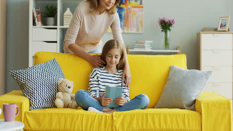 blonde cute girl sitting on yellow couch at home and watching something on tablet, then her mother approachs and hugs her 1