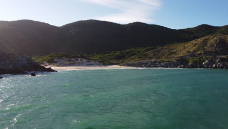 The-sun-shines-behind-the-mountains-on-the-horizon,-turquoise-waters-of-South-China-Sea-splash-near-the-beach-with-white-sand