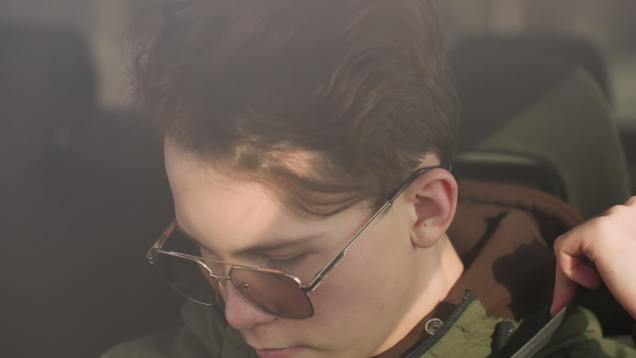 taxi driver in green winter jacket and glasses sitting inside vehicle with sunlight casting across face, preparing to fasten seatbelt, warm natural lighting visible through windshield reflection