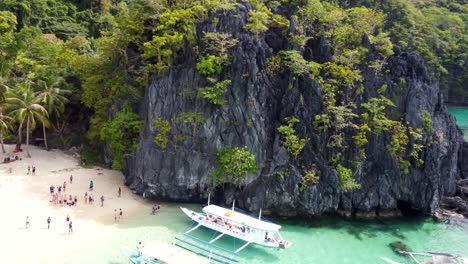 aerial: filipino outrigger tour boats loading and unloading tourists on tropical seven commandos beach, turquoise clear water on white sand, palm trees and lush green limestone mountains