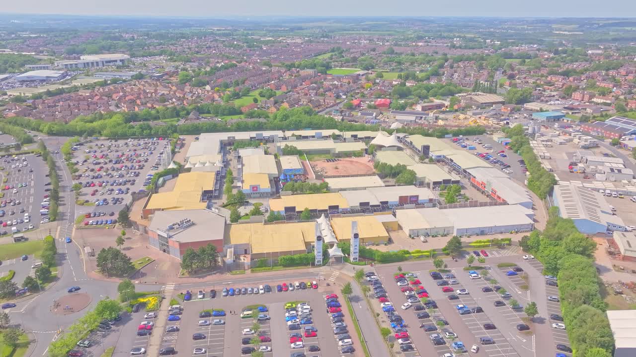 Slow aerial drone glide above Junction 32 outlet shopping village in Castleford, UK, showing layout, parking area, surrounding streets and distant townscape, summer daylight, slow motion
