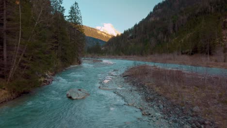 aerial flight along a scenic mountain river with fresh blue water in the bavarian austrian alps by sunshine , flowing down a riverbed along trees, rocks, forest and hills seen from above by drone