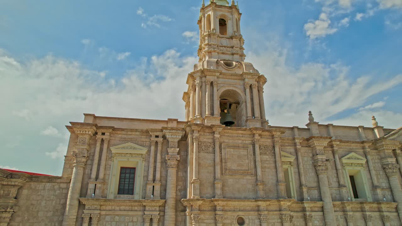 Upward drone pan of Arequipa Cathedral's left tower on a cloudy day with the sun illuminating the tower, a mix of sun and clouds