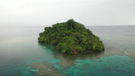 drone shot backwards of a small, lush and green island with a small, white boat next to it