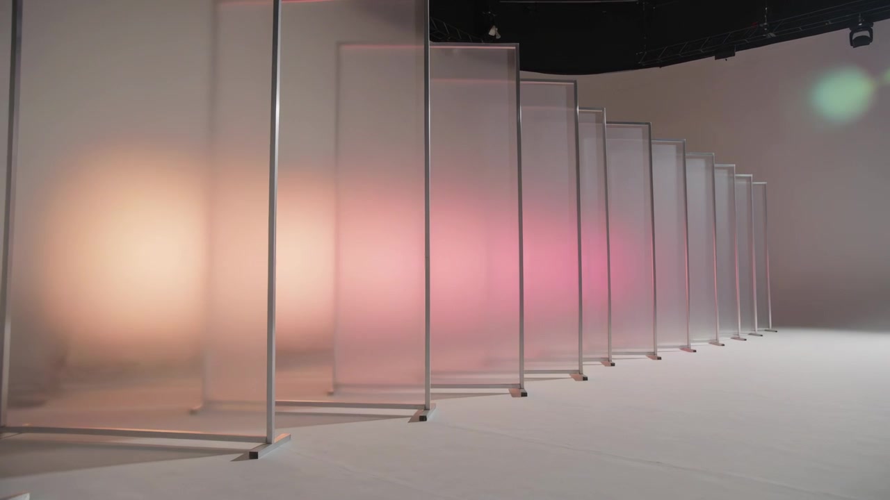 Luci che si accendono, telecamera che si sposta a destra mostrando pannelli con cornice metallica in studio, con luce rosa che crea profondità.