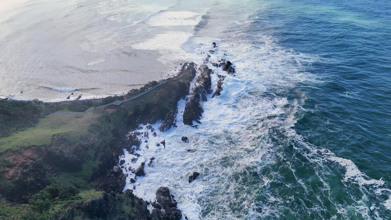 Drone footage captures the rugged Byron Bay coastline with crashing waves and lush greenery under natural daylight