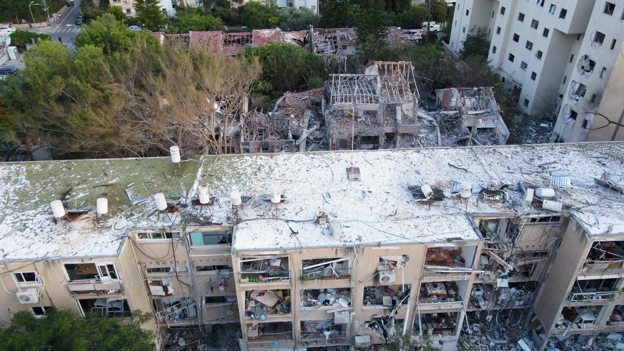 Drone flies over dusty rooftop in Tel Aviv, passing a blast-damaged tree and revealing a building reduced to rubble.