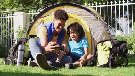 Happy-biracial-man-and-his-son-using-smartphone-in-garden