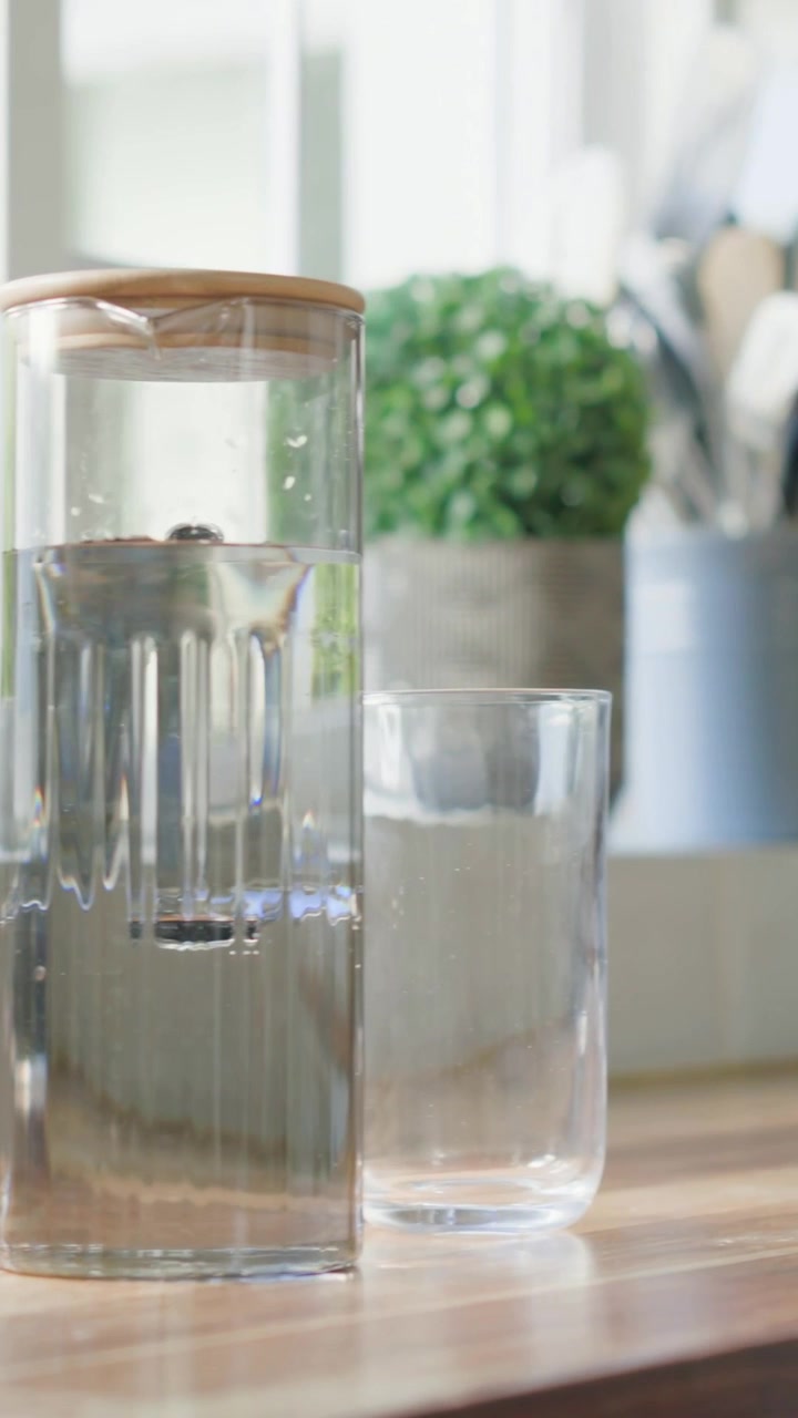 Vertikales Video: Sprudelnde Infusionskaraffe setzt Perlen auf Küchenarbeitsplatte durch Sonnenwärme frei, daneben ein Trinkglas.