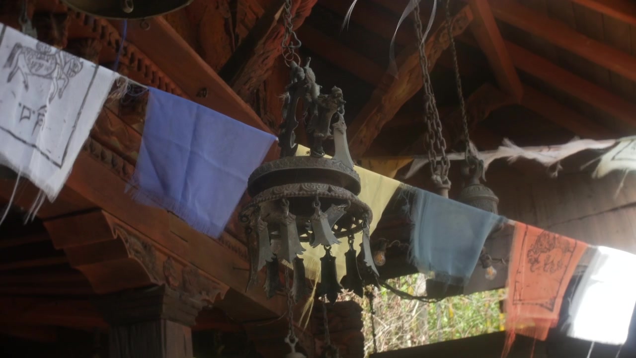 Campana appesa alle grondaie intagliate in legno di un tempio accanto a bandiere di preghiera colorate. La vista ravvicinata mostra catene di metallo, travi rustiche e una luce diurna soffusa in un cortile culturale.