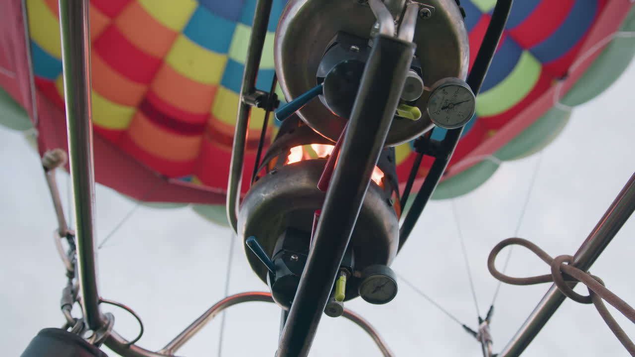 Close up gloved hand operating metal burner valve under vibrant balloon envelope panels, pilot controlling flame intensity via levers and pressure gauge against clear sky backdrop over green field