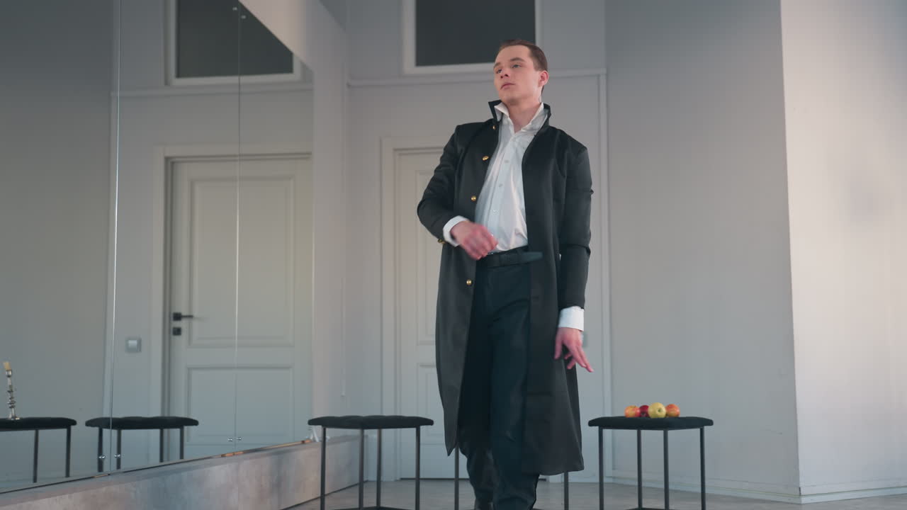 student in black coat and white shirt performs dramatic solo dance across studio floor, moving with intent past stools holding apples and candle stand, framed by mirror
