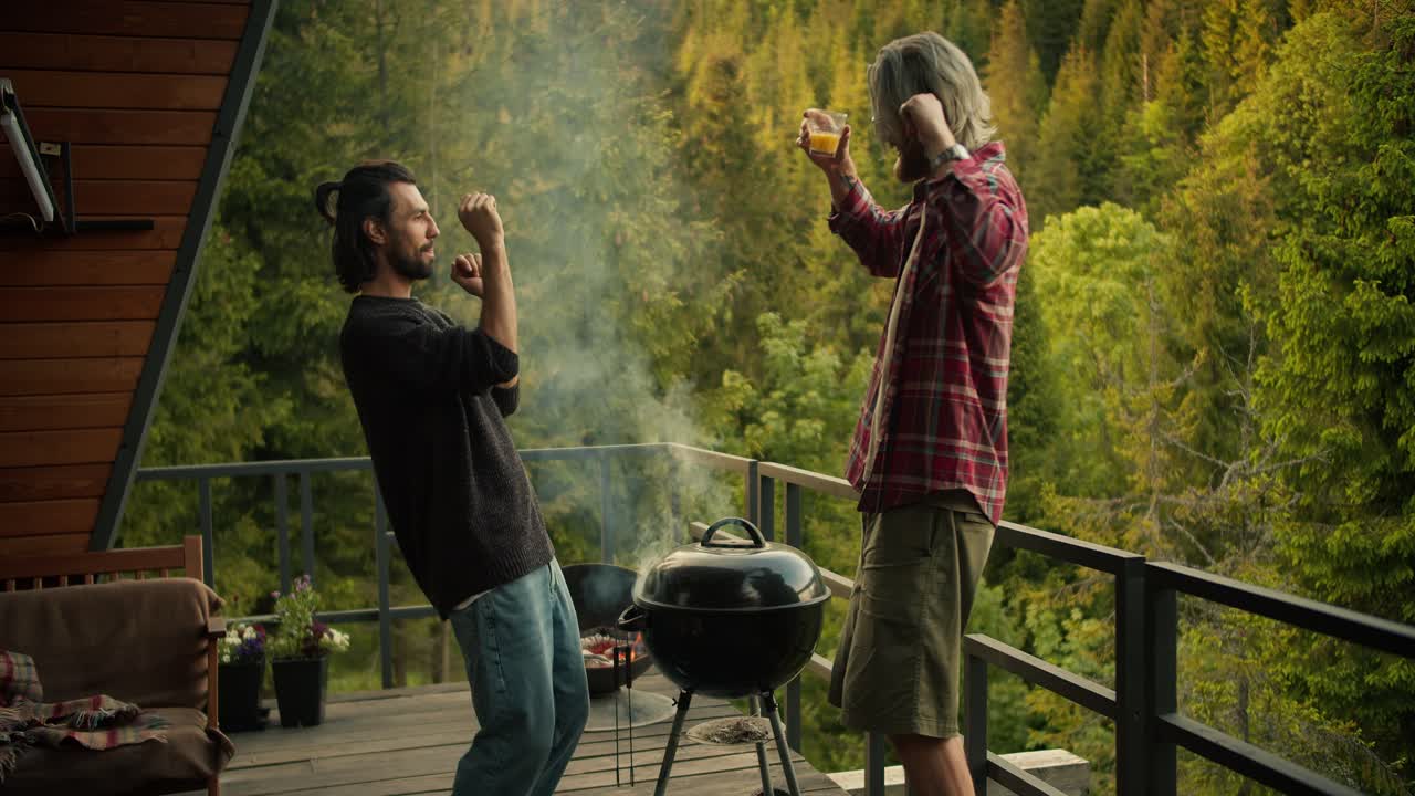 Due ragazzi ballano e si divertono mentre cucinano alla griglia su un ampio balcone con vista su conifere ghiacciate e montagne. Divertimento da picnic, tempo libero felice