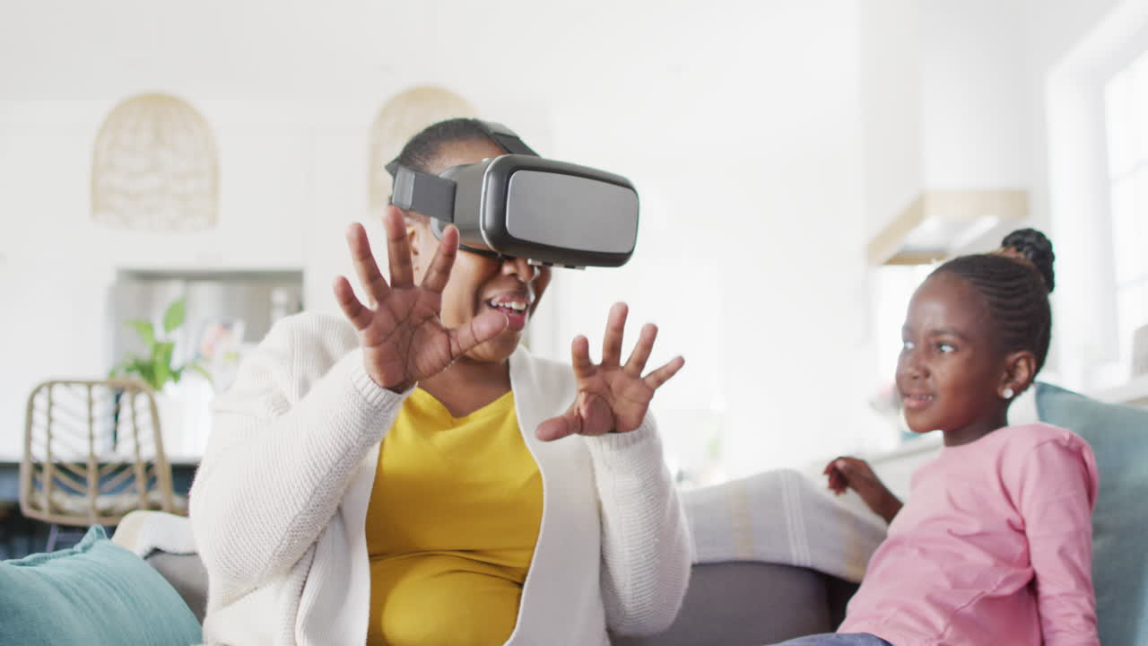 Excited african american grandmother using vr headset sitting with granddaughter at home, copy space
