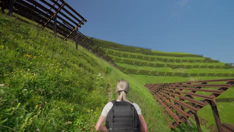a young woman hikes near snow barriers on a green hillside under a bright blue sky