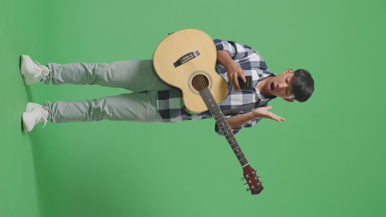 Adolescente asiatico a figura intera con la chitarra che usa lo smartphone e dice 'Wow' mentre è in piedi su sfondo a schermo verde