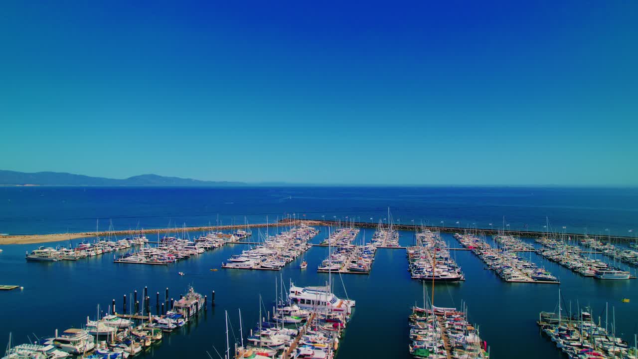 Santa Barbara Harbor showcasing rows of sailboats and yachts docked under a vibrant blue sky and north pacific ocean.