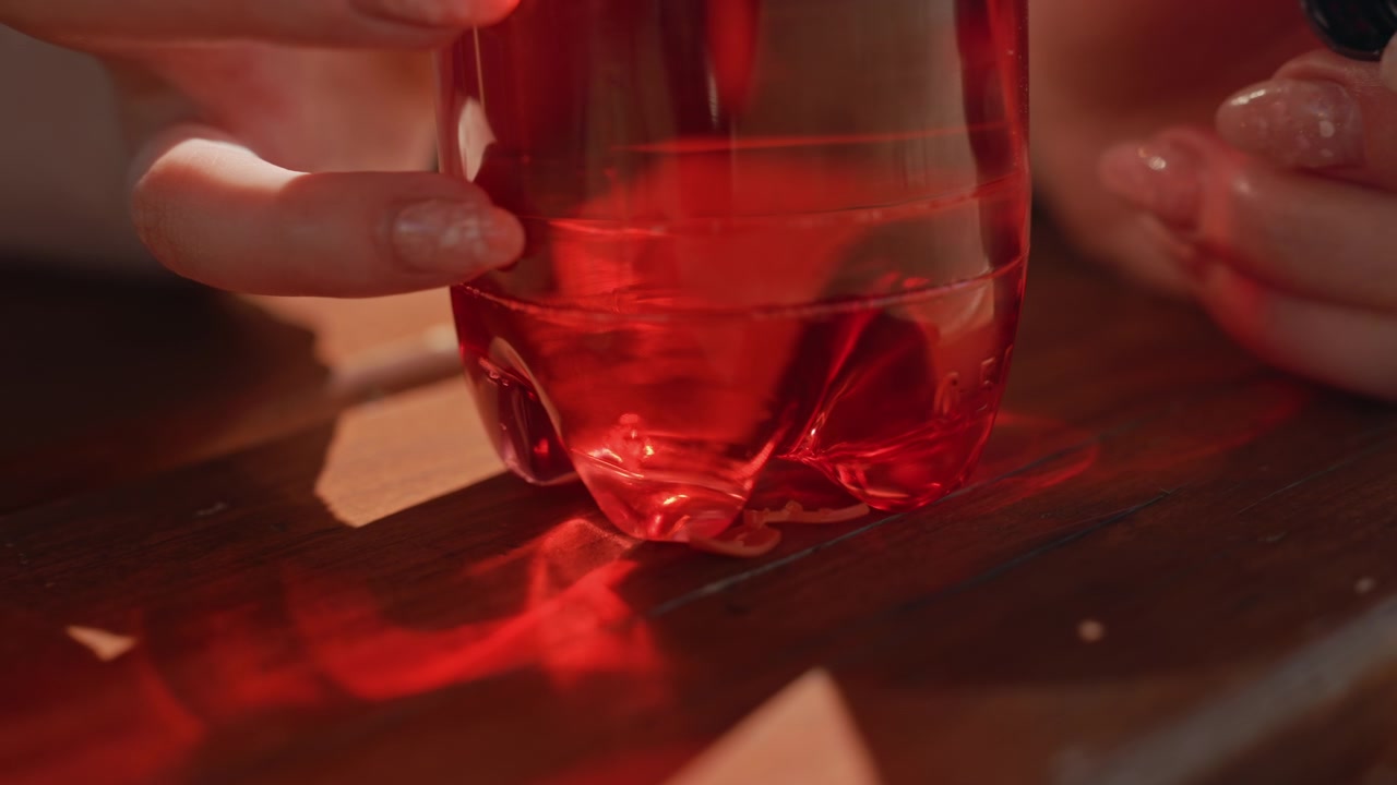 Mani di donna manipolano una bottiglia di plastica trasparente piena di liquido rosso, creando riflessi e rifrazioni su una superficie di legno. Una vista ravvicinata enfatizza i dettagli della texture della pelle e delle unghie dipinte.